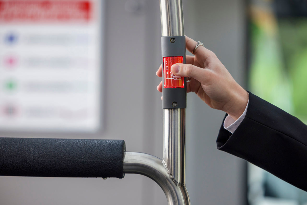 Eine Hand drückt im Inneren eines Stadtbusses auf den roten Halteknopf, der an einer senkrechten Metallstange befestigt ist.
