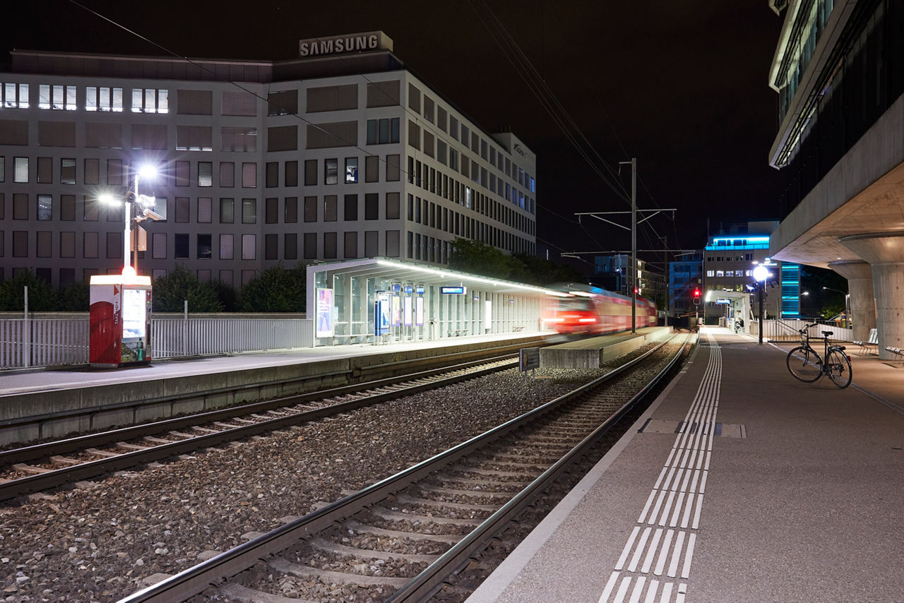 "Zwei Arbeiter in orangefarbener Sicherheitskleidung und Gurten führen Wartungsarbeiten an einem Zugdach in einer modernen Serviceanlage durch. Einer sitzt und arbeitet an einer elektrischen Komponente, während der andere über die geöffnete Klappe ins Innere blickt. Beide sind mit Sicherheitsleinen gesichert. Der Zug trägt das Logo der Schweizerischen Bundesbahnen (SBB).