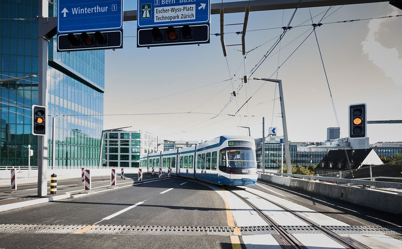 Cobra-Tram der VBZ in Zürich auf der Hardbrücke, umgeben von Glasfassaden und klarer Verkehrsführung mit Hinweisschildern nach Winterthur, Bern und Basel.
