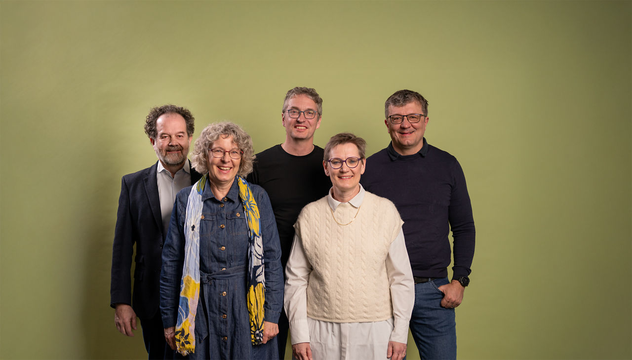 Eine Gruppe von fünf Erwachsenen steht vor einem hellgrünen Hintergrund und lächelt in die Kamera. Zwei Frauen stehen vorne, eine trägt ein langes Jeanskleid mit buntem Schal, die andere ein weisses Hemd mit cremefarbener Strickweste. Dahinter stehen drei Männer, alle mit Brille; sie tragen legere bis halbformelle Kleidung. Die Stimmung ist freundlich und entspannt.