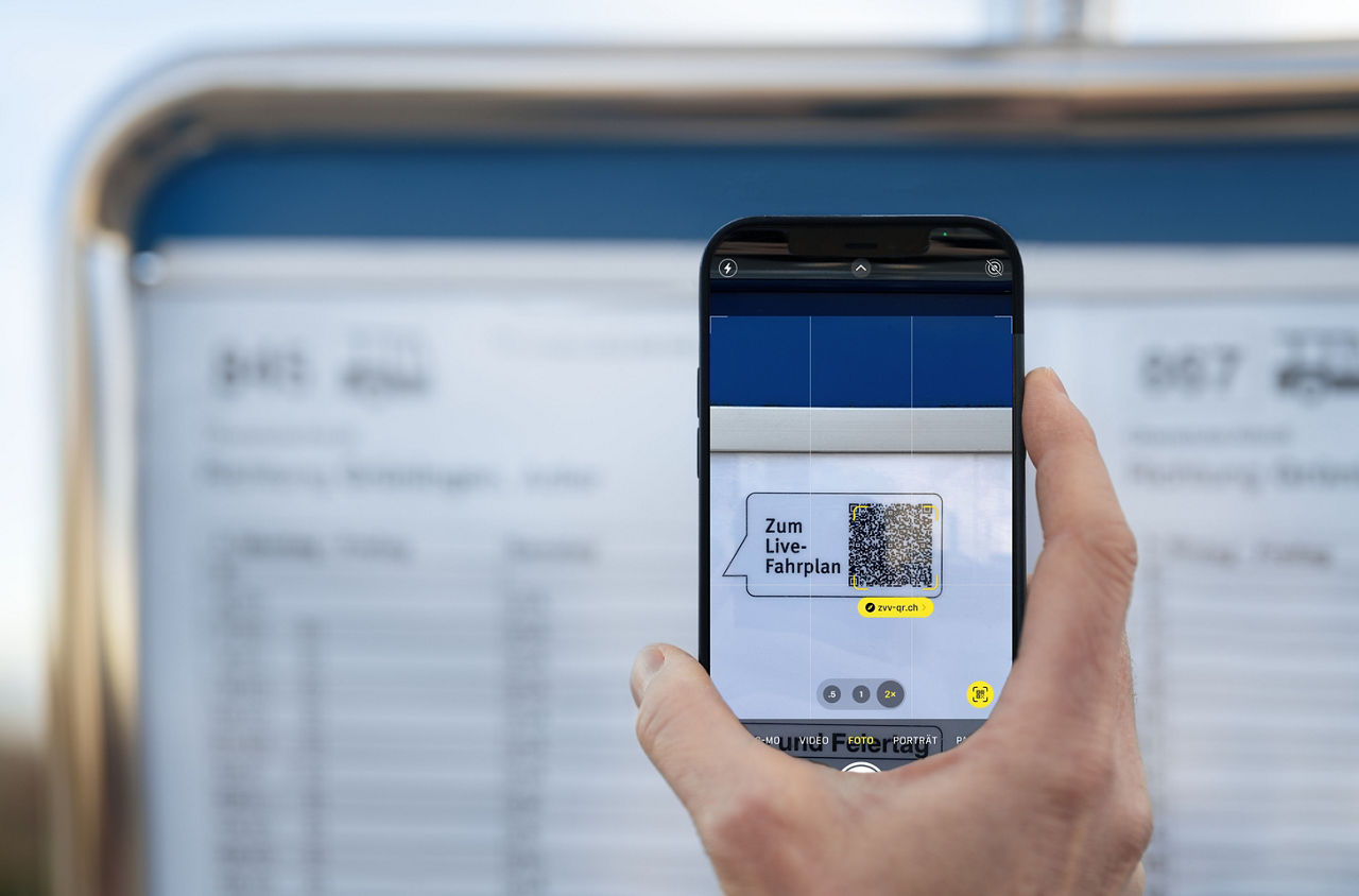 Eine Hand hält ein Smartphone, das einen QR-Code auf einem Haltestellenfahrplan scannt. Auf dem Smartphone-Bildschirm ist der QR-Code mit dem Text „Zum Live-Fahrplan“ und der URL „zvv-qr.ch“ sichtbar. Im Hintergrund ist der Haltestellenfahrplan unscharf zu erkennen.