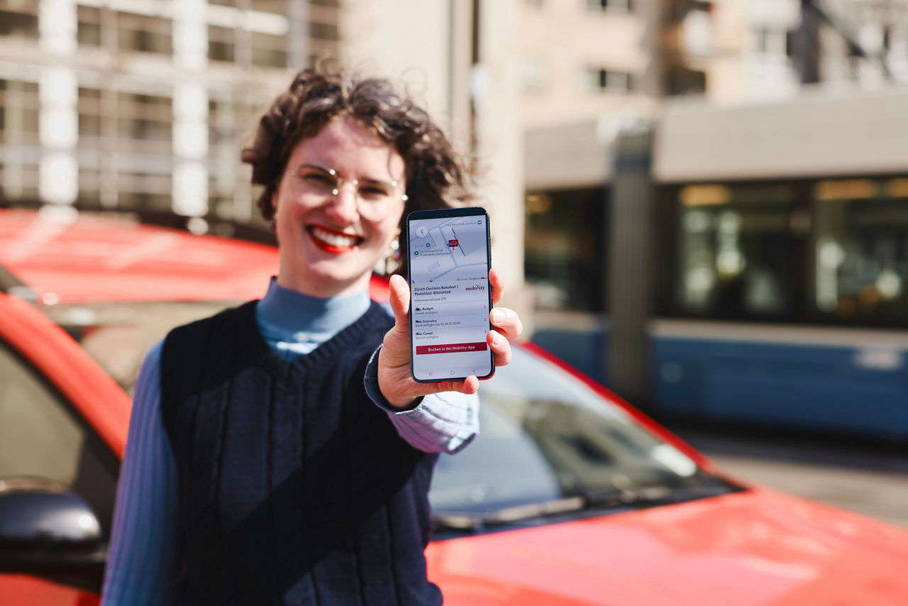 Eine lachende Frau steht vor einem roten Mobility-Auto und hält ein Smartphone in die Kamera. Auf dem Bildschirm ist die ZVV-App geöffnet, in der ein Mobility-Standort in Zürich angezeigt wird. Im Hintergrund ist ein blau-weisser Zürcher Tram zu sehen. Das Bild zeigt die Integration von Mobility in die ZVV-App, mit der Nutzer direkt über die App ein Mobility-Auto finden können.
