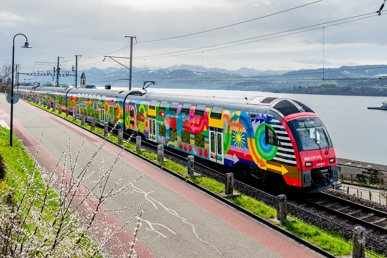 Ein farbenfroh bemalter Doppelstockzug der SBB (Zugnummer 514 015) fährt entlang eines Sees (Zürichsee) mit schneebedeckten Alpen im Hintergrund. Die auffällige Bemalung zeigt abstrakte Muster (ZVV Kampagne Gemeinsam Vorwärts)und leuchtende Farben. Der Zug fährt auf Gleisen parallel zu einem Rad- und Fussweg, im Vordergrund sind blühende Zweige zu sehen