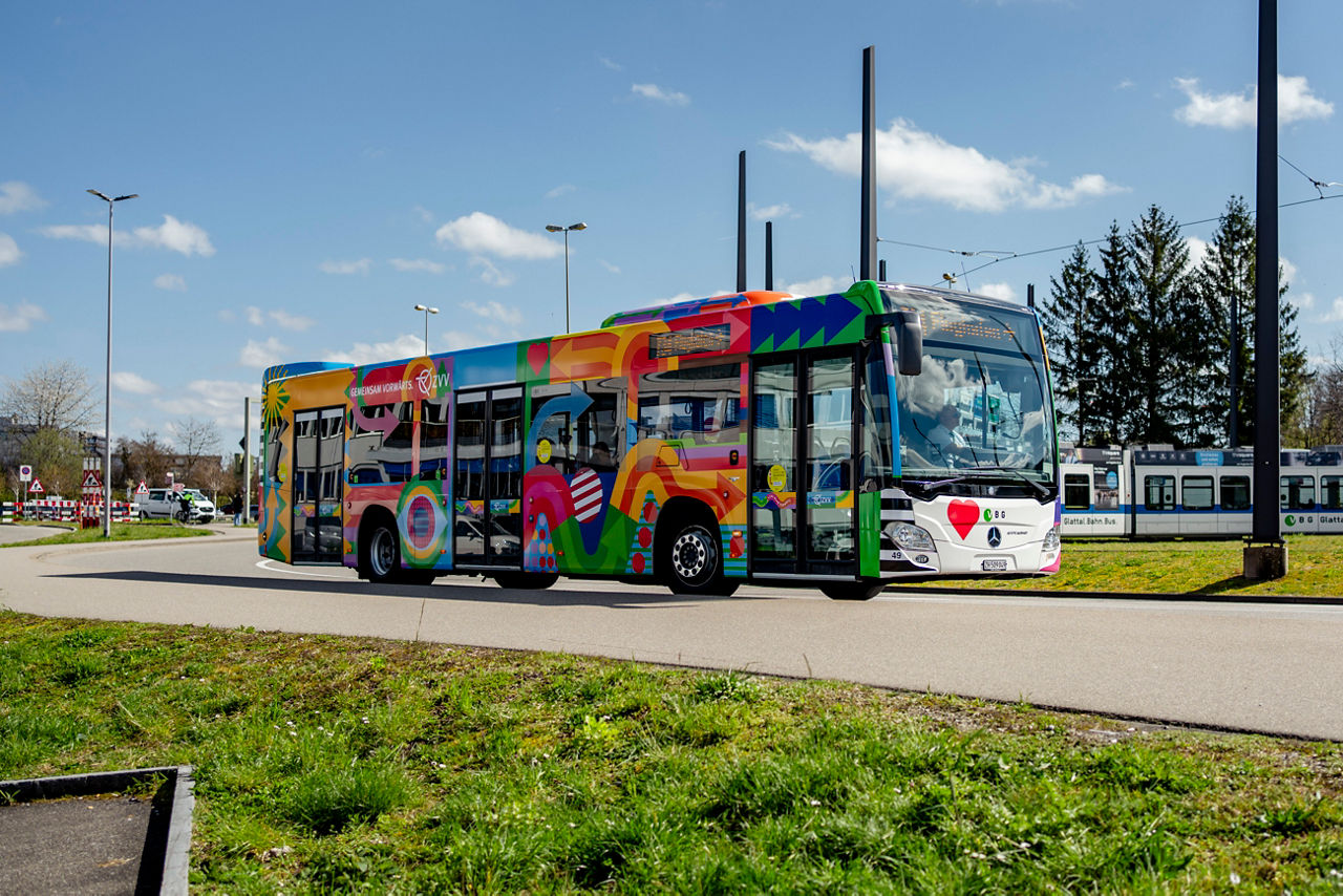 Ein moderner Linienbus der VBG (Verkehrsbetriebe Glattal) mit einer auffälligen, farbenfrohen Vollbemalung steht auf einer Strasse an einem sonnigen Tag. Die Busgestaltung zeigt bunte, abstrakte Muster mit Pfeilen, Herzen, Kreisen und Wellen in kräftigen Farben wie Rot, Grün, Blau, Gelb und Rosa. Auf der Seite des Busses ist der Schriftzug „GEMEINSAM VORWÄRTS“ zusammen mit dem ZVV-Logo (Zürcher Verkehrsverbund) zu sehen. Im Hintergrund sind ein Tram und Bäume sichtbar, darüber ein klarer blauer Himmel mit einigen Wolken. Die Szene vermittelt einen lebendigen, positiven Eindruck.